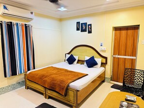Habitación de hotel con una cama, cabecera de madera, manta marrón, aire acondicionado de pared, ventana con persianas y puerta con panel de madera.