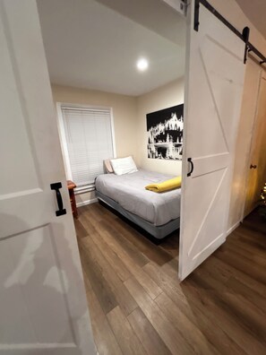 Een slaapkamer met een bed, een raam met blinds, een aan de muur hangend kunstwerk en een houten vloer.