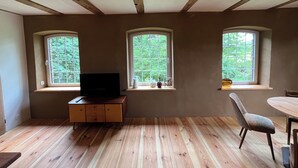 A room with wooden beams, a TV on a wooden cabinet, and three windows with greenery outside.