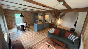 A rustic interior with exposed wooden beams, a dining area with a round table and chairs, a kitchen with blue cabinets, and a green sofa with red cushions.