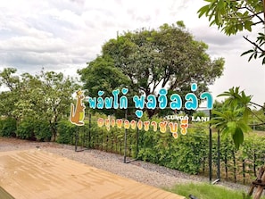 Un letrero con texto tailandés e ilustración de un canguro, rodeado de exuberante vegetación y una terraza de madera.