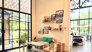 Una habitación amplia con ventanas grandes, una mesa de madera, una silla de mimbre, un sofá con cojines verdes y estantes con marcos y plantas en macetas.