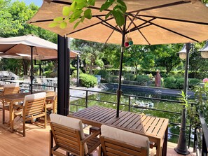 An outdoor patio with wooden tables and chairs, umbrellas, and a view of a water feature. - Plumgoland (Pak Tho)