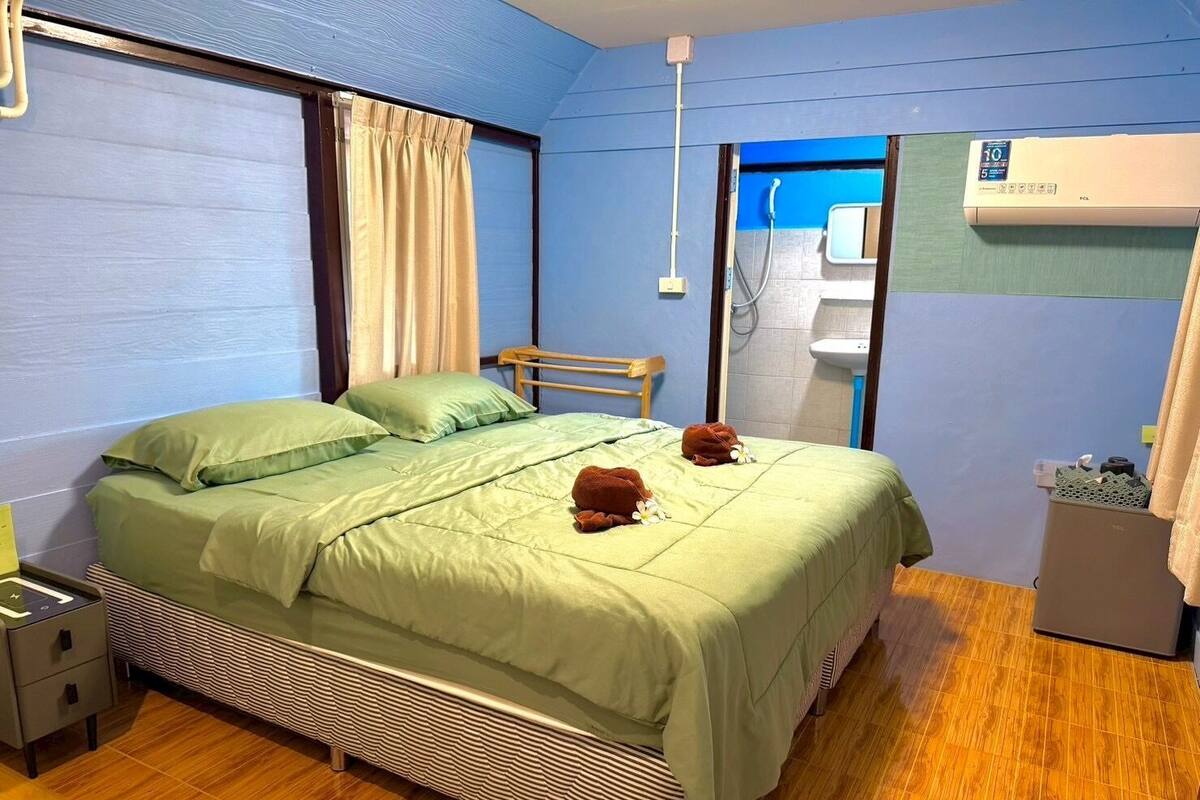 A single bed with a green bedspread, a wooden headboard, a bedside table with a lamp, and a window with curtains.