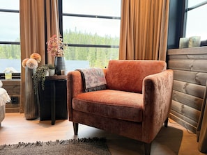 A cozy room with a plush orange armchair, a wooden sideboard, and a view of trees through large windows. - New cabin with activities and fire pit near Voss (Voss)