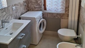 Une petite salle de bain comprenant un lavabo, un lave-linge, des toilettes et une fenêtre agrémentée de rideaux à motifs.