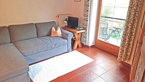 Een woonkamer met een grijze bank, een klein houten tafeltje met een lamp, een televisie en uitzicht buiten door een raam met gordijnen.