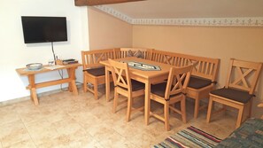 Een eethoek met een houten tafel en stoelen, een aan de muur bevestigde televisie en een vloerkleed met een patroon.