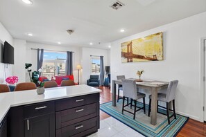 A modern kitchen with a central island, dining area with chairs, a painting on the wall, and a view through the window.