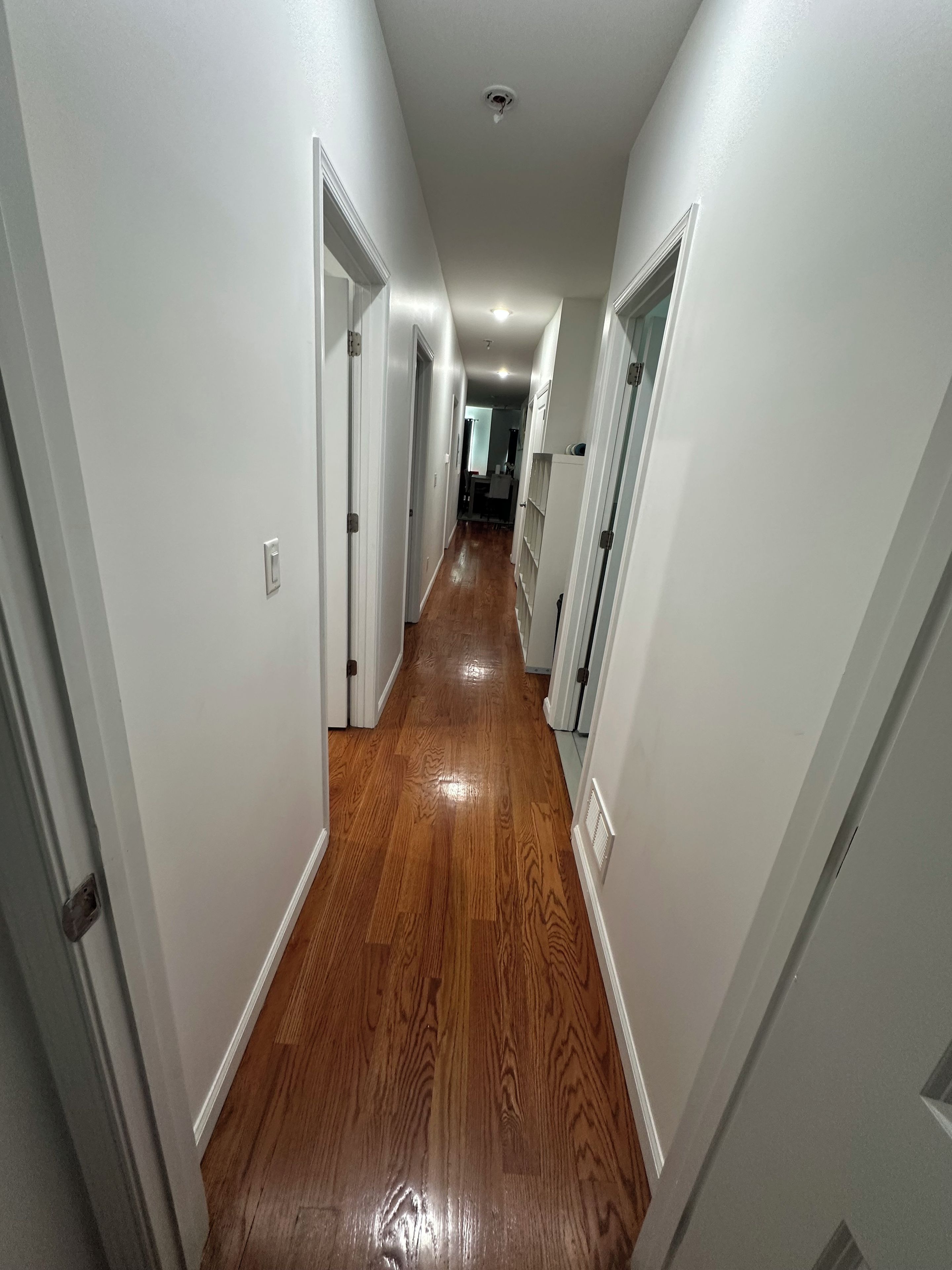 A hallway with white walls and wooden flooring leading to a room with a window.