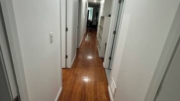 A hallway with white walls and wooden flooring leading to a room with a window.