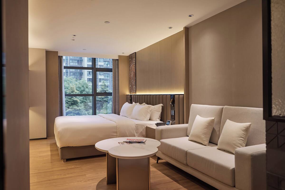 A modern hotel room with a large bed, a sofa, a small round table, and a large window.