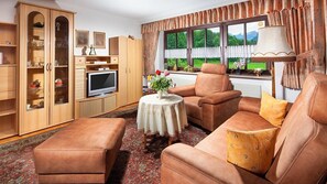 Ein Wohnzimmer mit einer Couch, einem Hocker, einem Fernseher, einem Holzschrank und einem Fenster mit Blick auf Grünfläche.