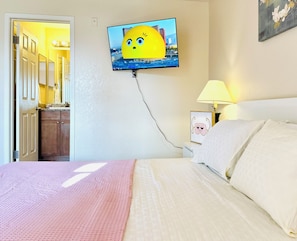 Un dormitori amb un llit, un televisor penjat a la paret, una làmpada de nit i una porta que dona a un bany.