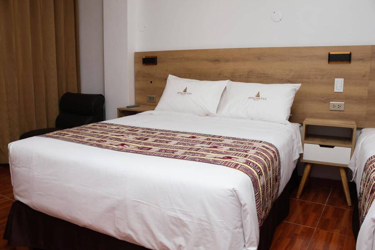 Une chambre d’hôtel avec une tête de lit en bois, un grand lit avec une literie à motifs, deux tables de chevet avec des tiroirs et une chaise.