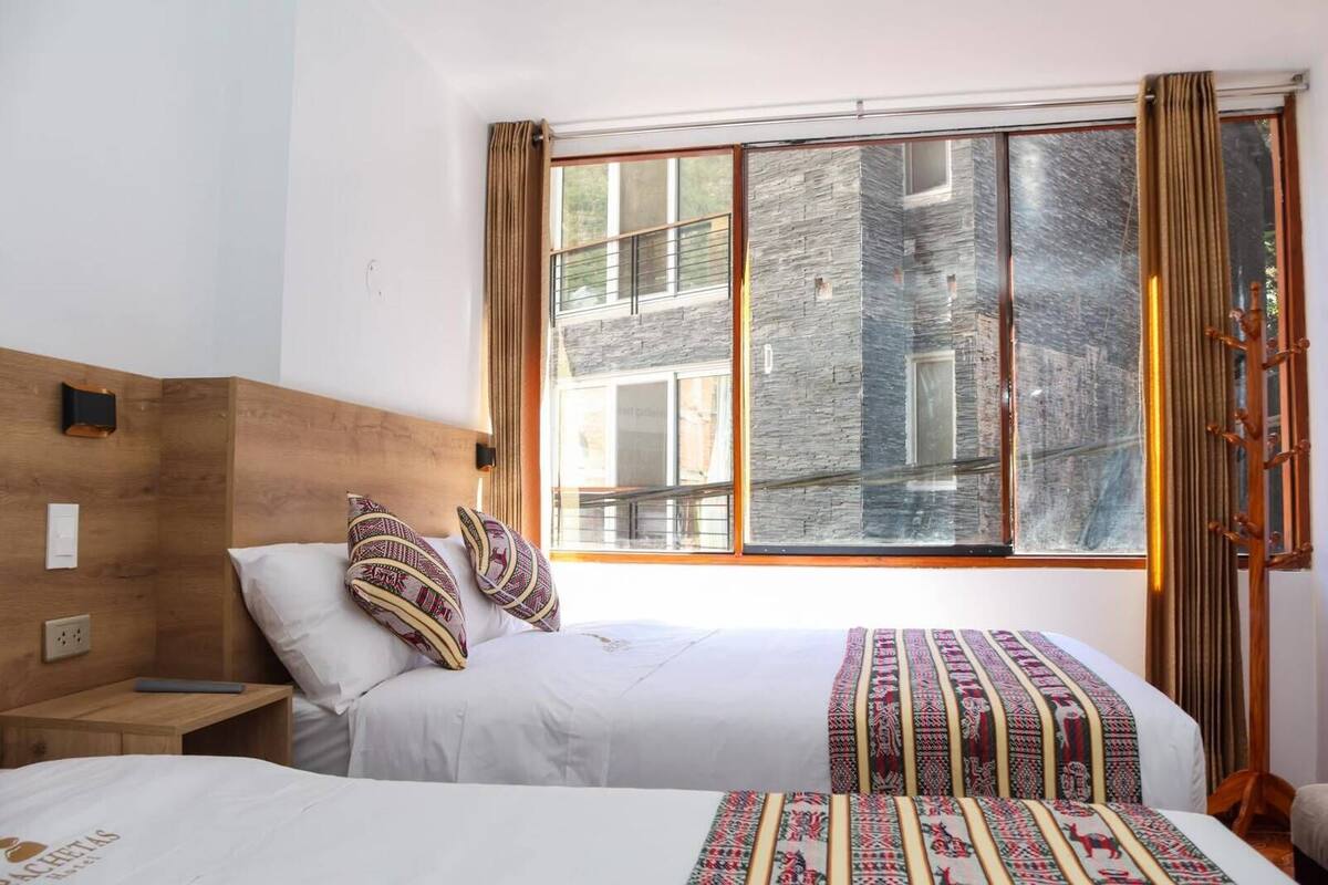 Une chambre d’hôtel avec une grande fenêtre, un lit avec une literie à motifs, une tête de lit en bois et une table de chevet avec une lampe.