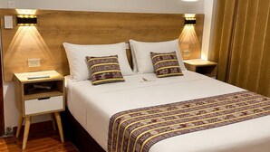 Habitación de hotel con cabecera de madera, dos mesitas de noche con lámparas, cama con ropa de cama estampada y suelo de madera.