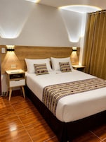 Habitación de hotel con cabecera de madera, dos mesitas de noche con lámparas, cama con ropa de cama estampada y suelo de madera.
