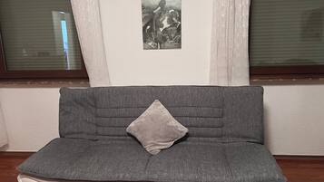 Ein graues Sofa mit einem Kissen, ein gerahmtes Bild an der Wand und ein Fenster mit Jalousien.