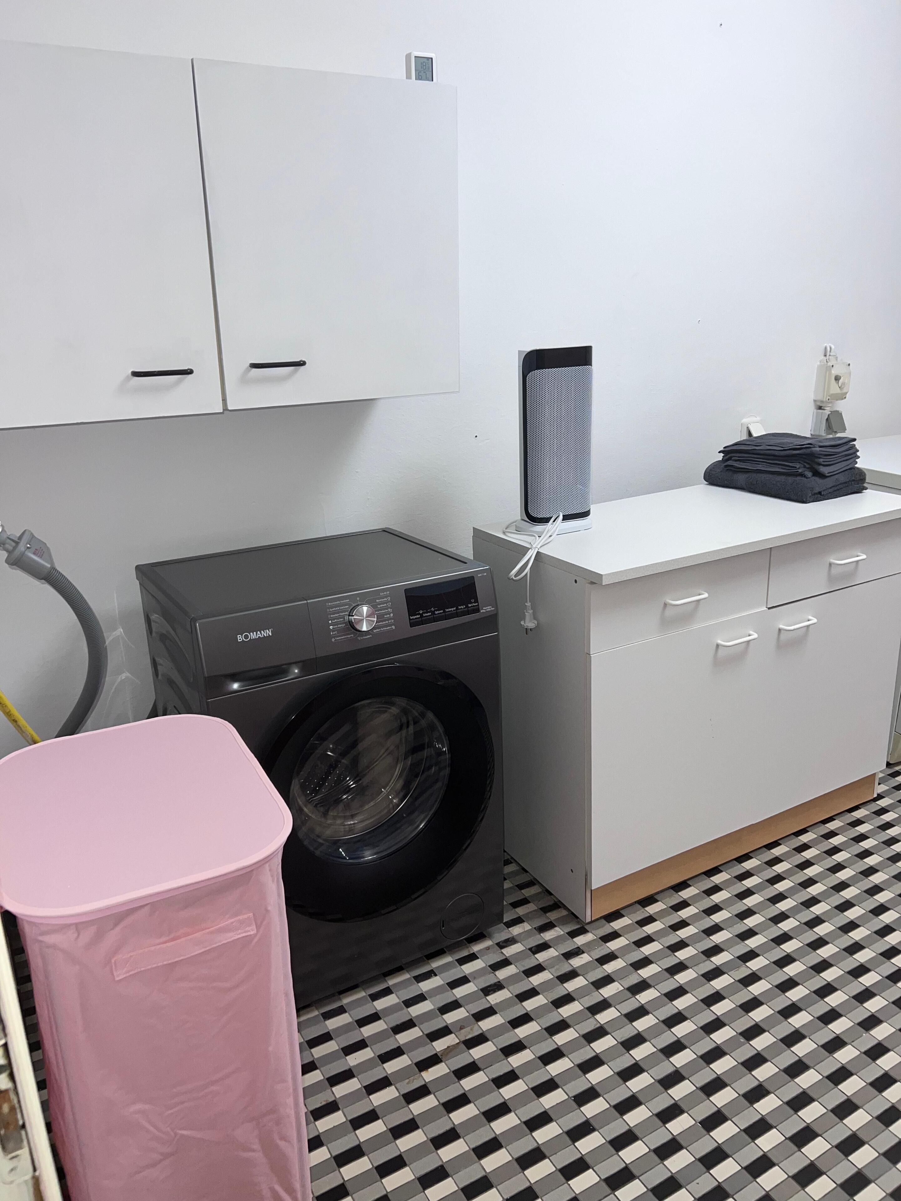 A laundry area with a washing machine, a dryer, a small table with a speaker, and a pink trash bin.