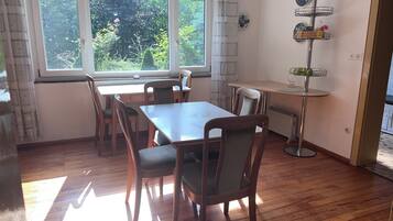 A room with a wooden table and chairs, a window with a view of greenery, a radiator, and a small shelf with items.