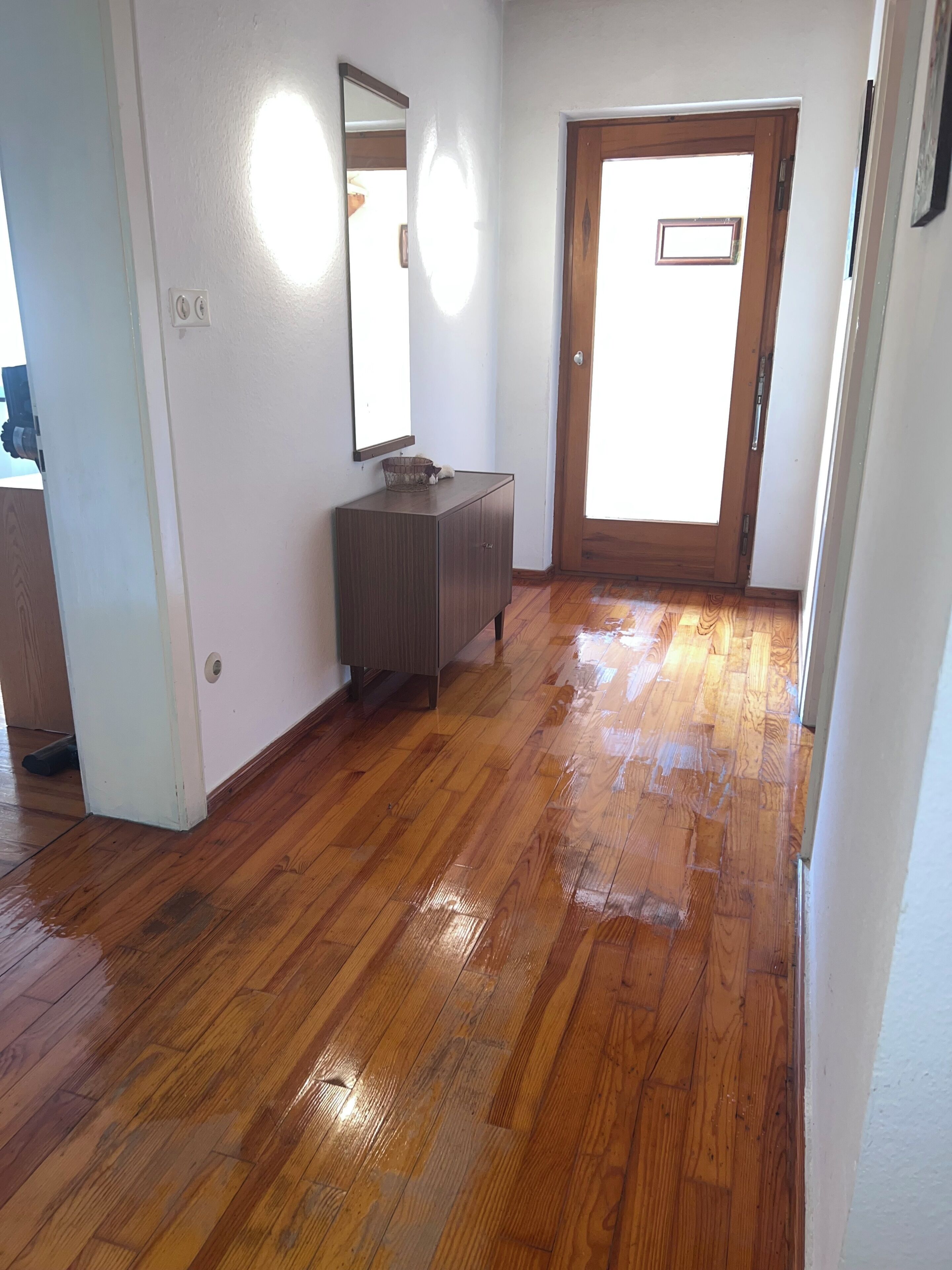 Una habitación con piso de madera, una puerta de madera y una pared blanca con un espejo.