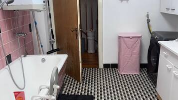 A bathroom with a bathtub, a shower, a toilet, a washing machine, and a pink trash bin.