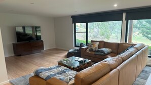 Un soggiorno moderno con un divano in pelle, un tavolino in vetro, una TV su un mobile in legno e ampie finestre con vista sul verde.