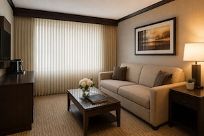 Ein Hotelzimmer mit einer Couch, einem Couchtisch mit Blumen, einer Lampe, einem gerahmten Bild an der Wand und einem Fernseher auf einem Schrank.