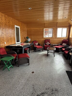 A room with wooden walls, a pool table, wicker chairs with red cushions, and a small table with a fan.
