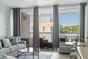 Ein modernes Wohnzimmer mit einer Couch, einem Couchtisch und Sesseln, ein Balkon mit Geländer und Blick auf Gebäude und Bäume.