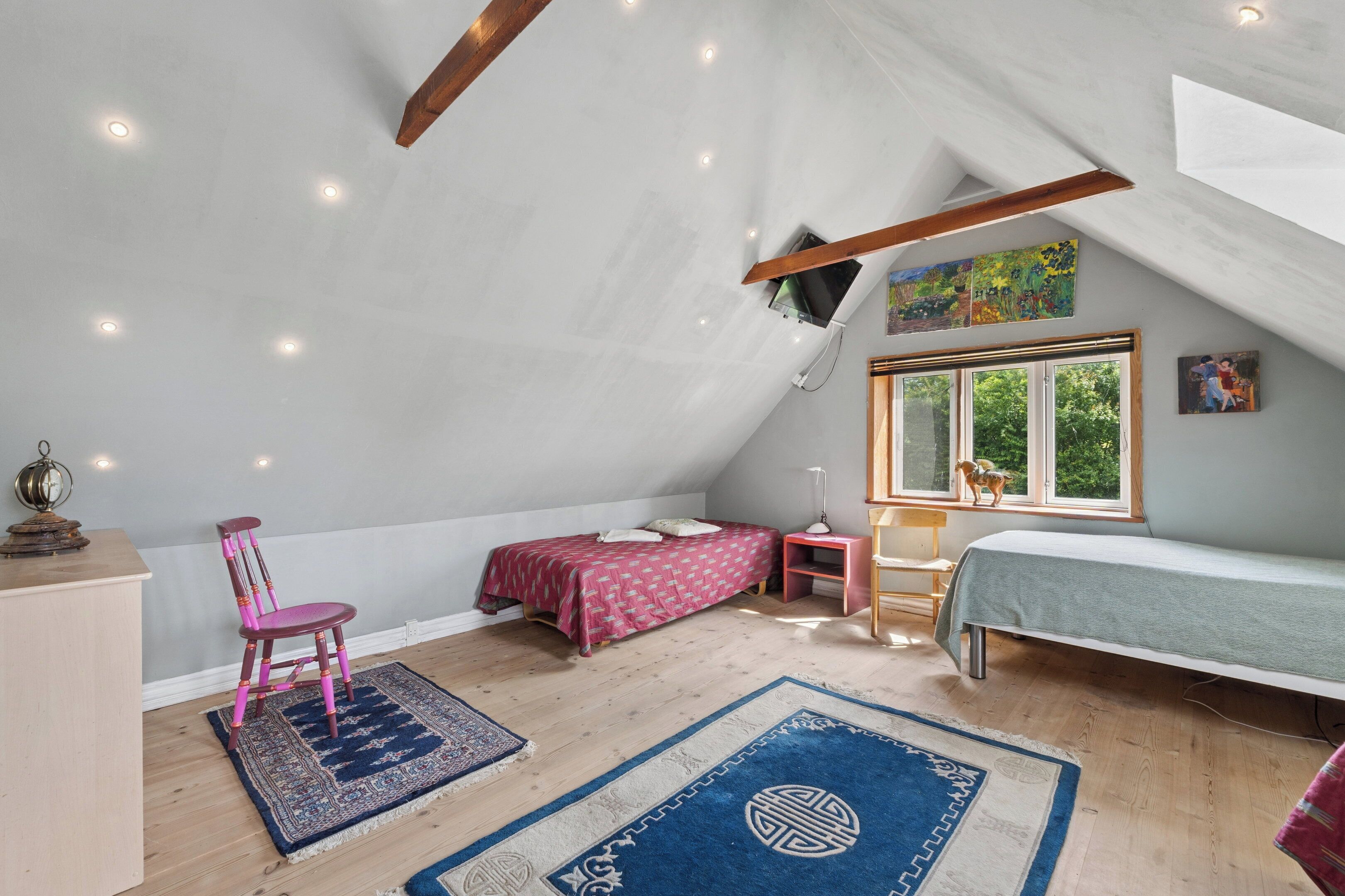 A bedroom with a sloped ceiling, a bed, a chair, a desk, and a window with a view of greenery.