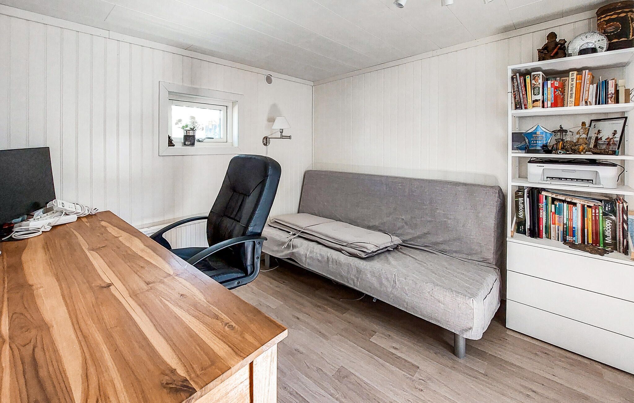 Een kamer met een houten bureau, een grijze bank, een boekenkast en een raam met uitzicht.