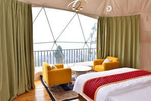 Una habitación con una cama, dos sillas amarillas, una mesa redonda pequeña y vistas amplias a través de grandes ventanales.