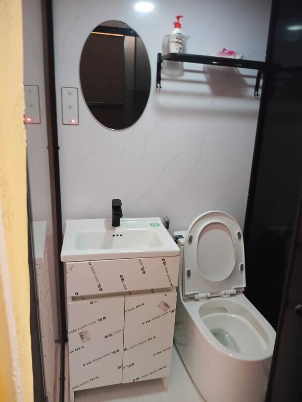 Un baño pequeño con inodoro, lavamanos, espejo y repisa con una botella.