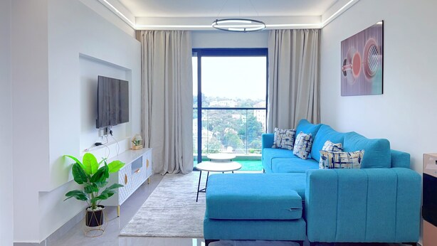 A modern living room with a blue sofa, a flat-screen TV, a potted plant, and a view through large windows. - Secure Lavington Apt w/ Pool, Gym & Fast WiFi – 1 Min Walk to Junction Mall (Nairobi)