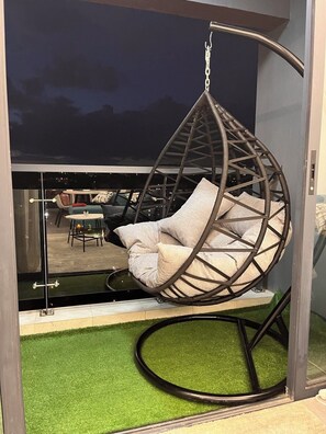 A hanging chair with cushions, a glass partition, and a view of an outdoor seating area. - Secure Lavington Apt w/ Pool, Gym & Fast WiFi – 1 Min Walk to Junction Mall (Nairobi)
