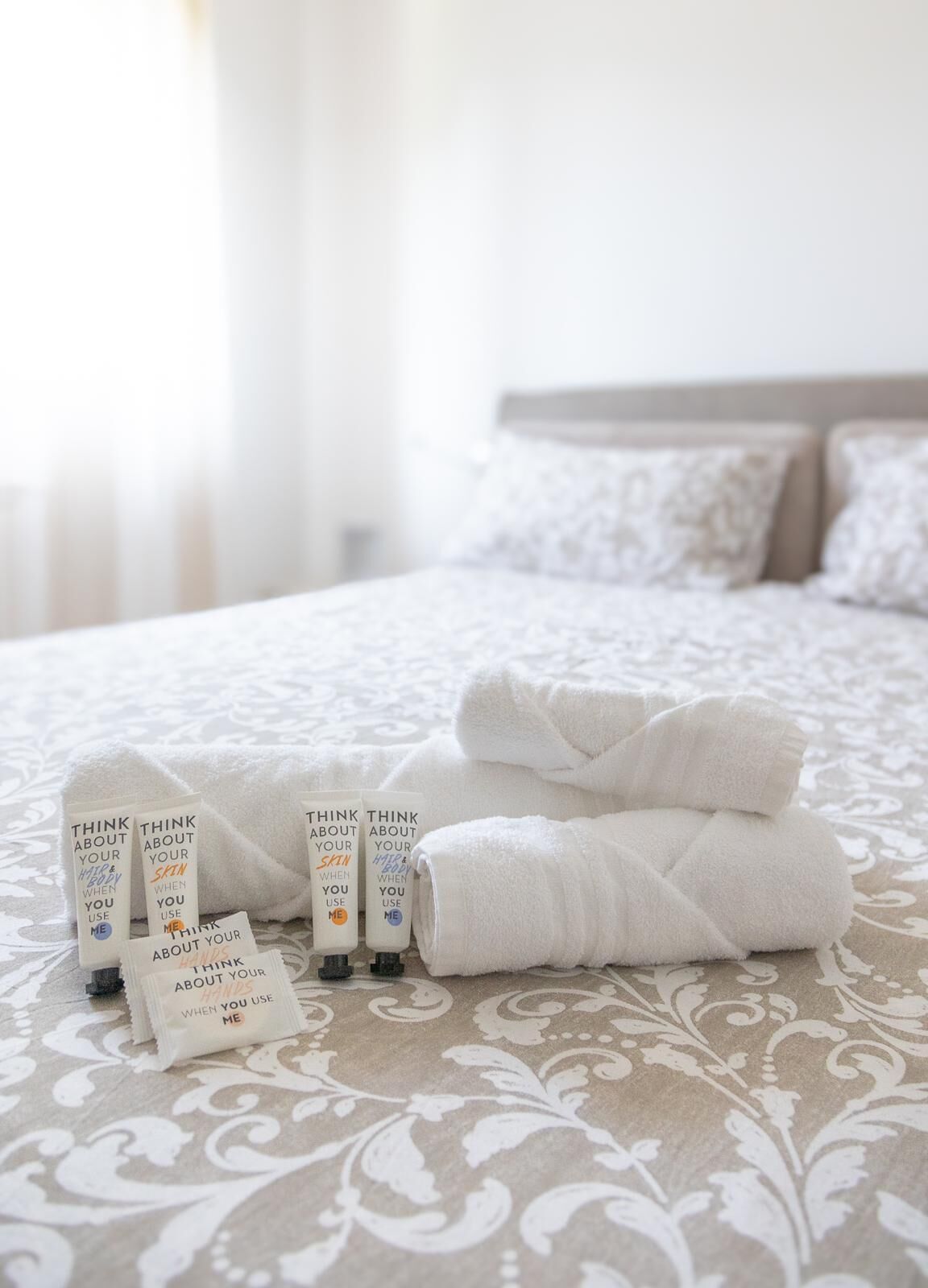 Un lit bien fait, avec des serviettes blanches pliées et plusieurs flacons de produits de soins pour la peau disposés sur une couvre-lit à motifs.