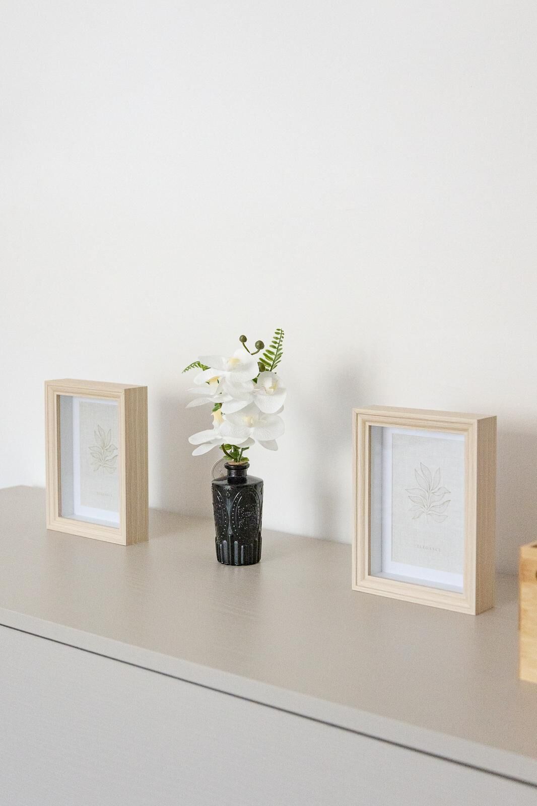 Ein Regal mit zwei gerahmten Pflanzenbildern und einer Vase mit weißen Blumen.