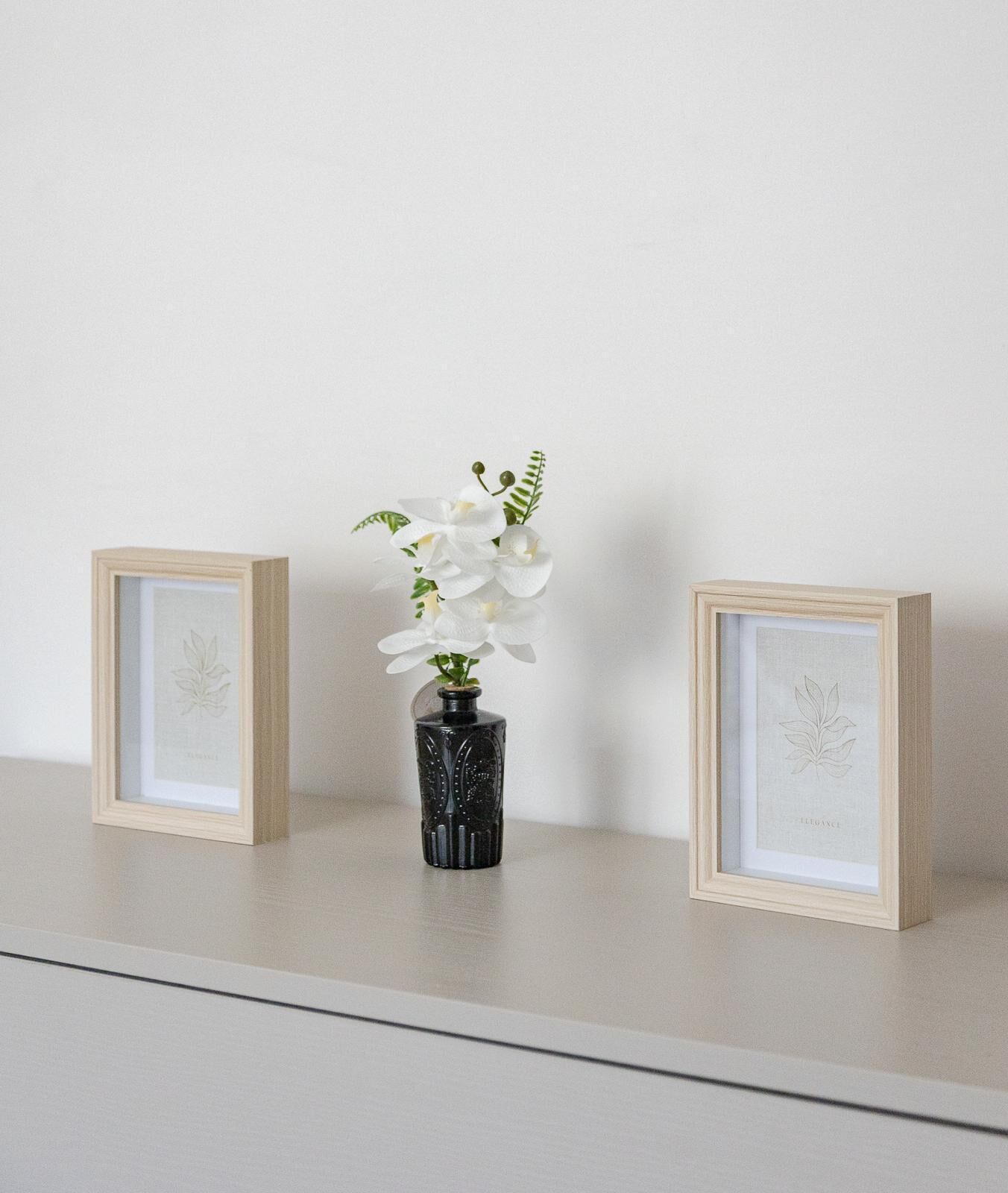 Een lijst met een botanische afbeelding op een plank, een zwarte vaas met witte bloemen, en nog een lijst.