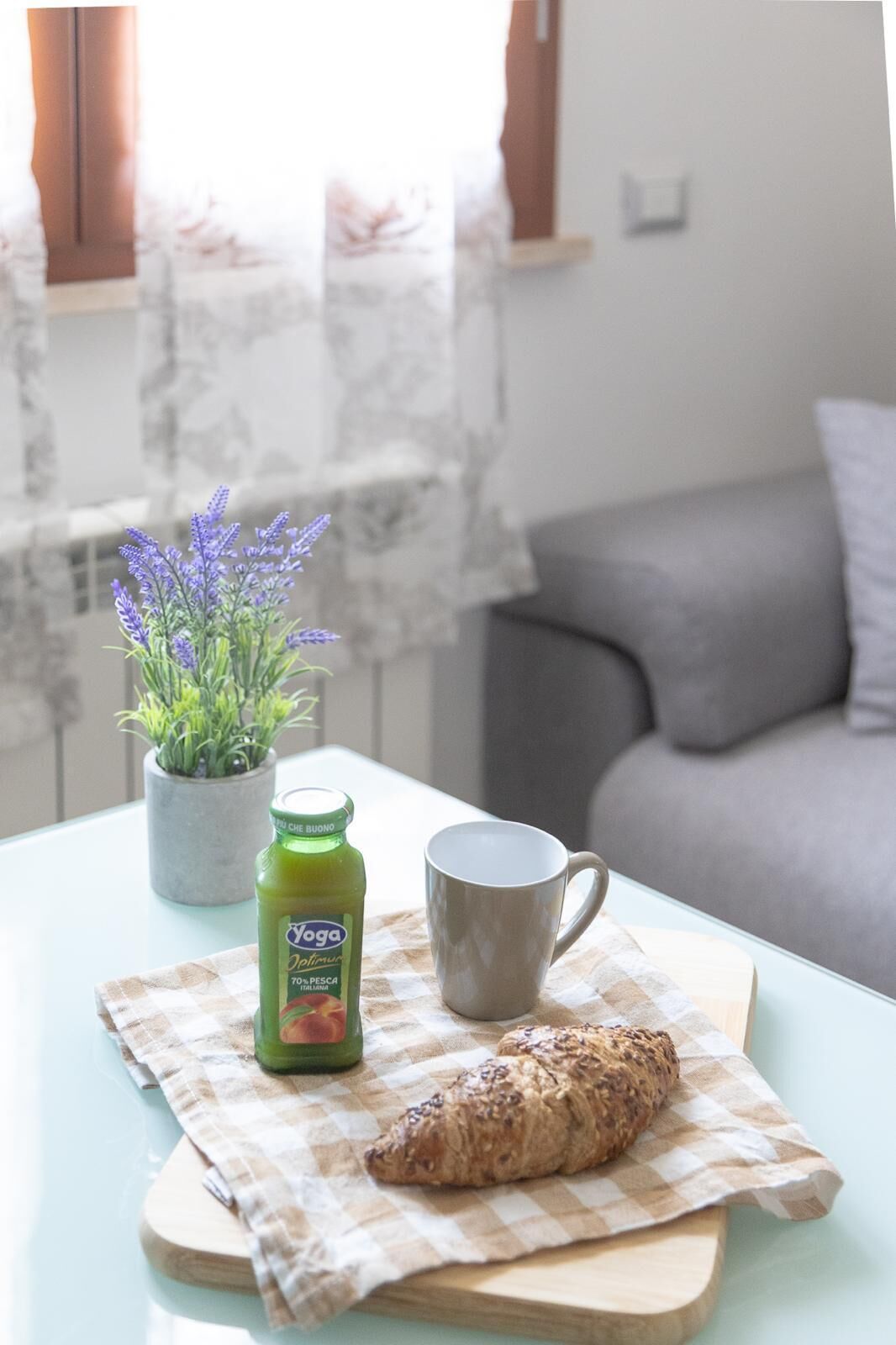 Uma mesa com toalha xadrez, uma garrafa de suco, uma caneca e um croissant sobre uma tábua de madeira.