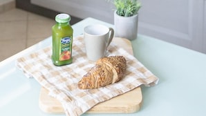 Een keuken met een fornuis, een kamerplant, een fles sap, een beker en een croissant op een tafel.