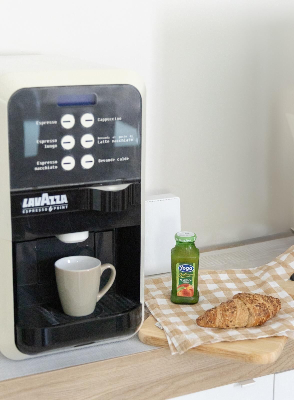 En kaffemaskine med kopholder, en juiceflaske og en croissant på en ternet serviet.