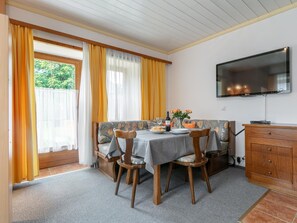 A dining area with a table set for two, a sofa, a television mounted on the wall, and a wooden cabinet.