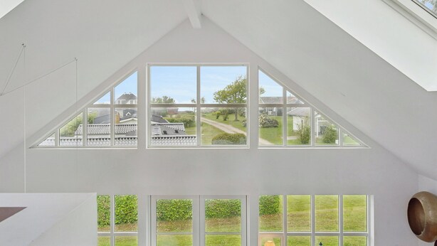 A room with a sloped ceiling, skylights, and large windows offering a view of a house and garden. - Scenic holiday home close to the water (Brenderup)