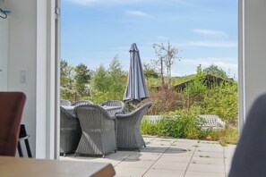 Eine Terrass mit Korbmöbeln und einem grauen Sonnenschirm, mit Blick auf einen Garten und ein Gebäude.