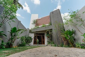 Un edificio moderno con fachada de hormigón, puerta de madera y un pequeño jardín con diversas plantas.