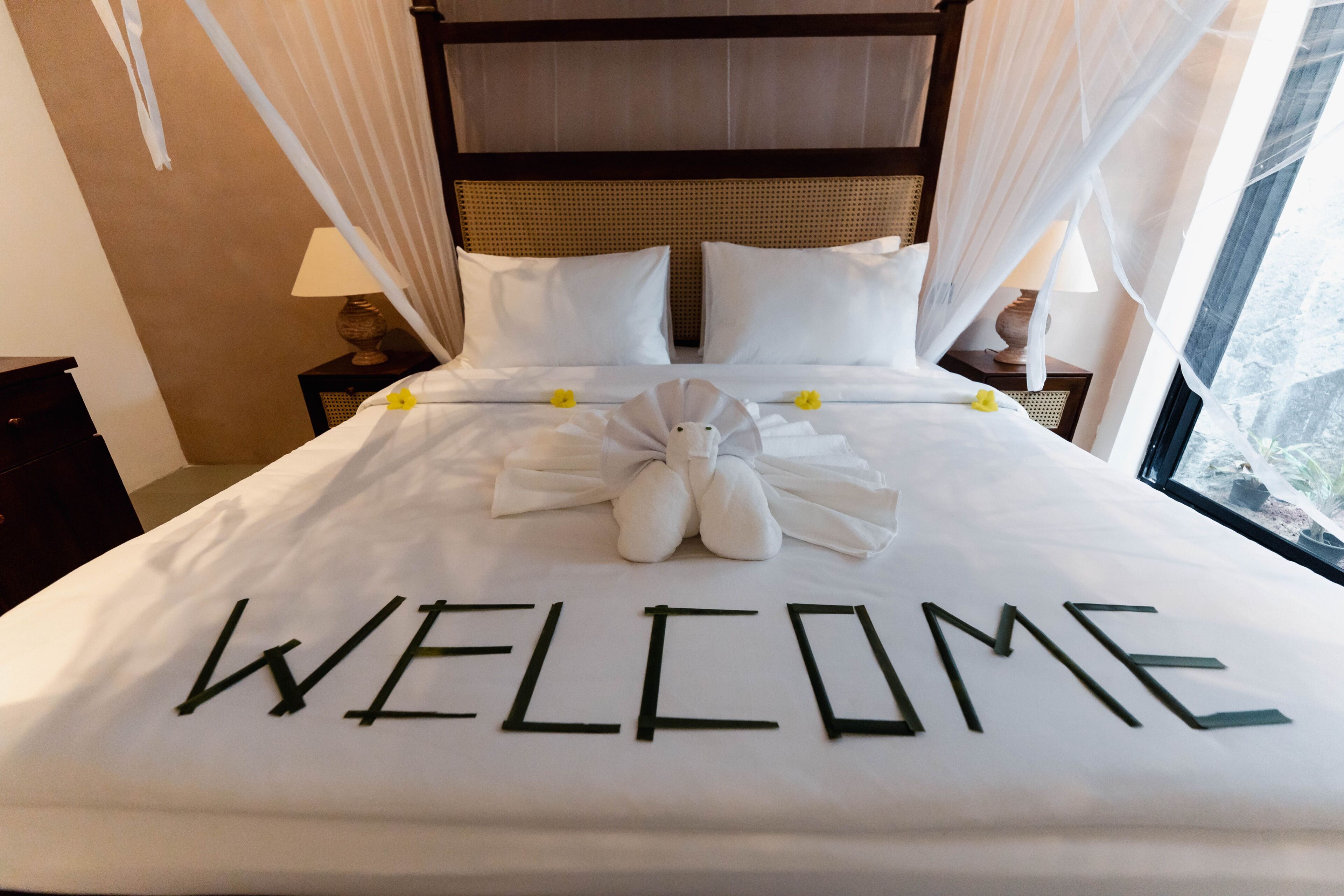 A neatly made bed with a 'WELCOME' sign and a plush toy, with a canopy and lamps on either side.