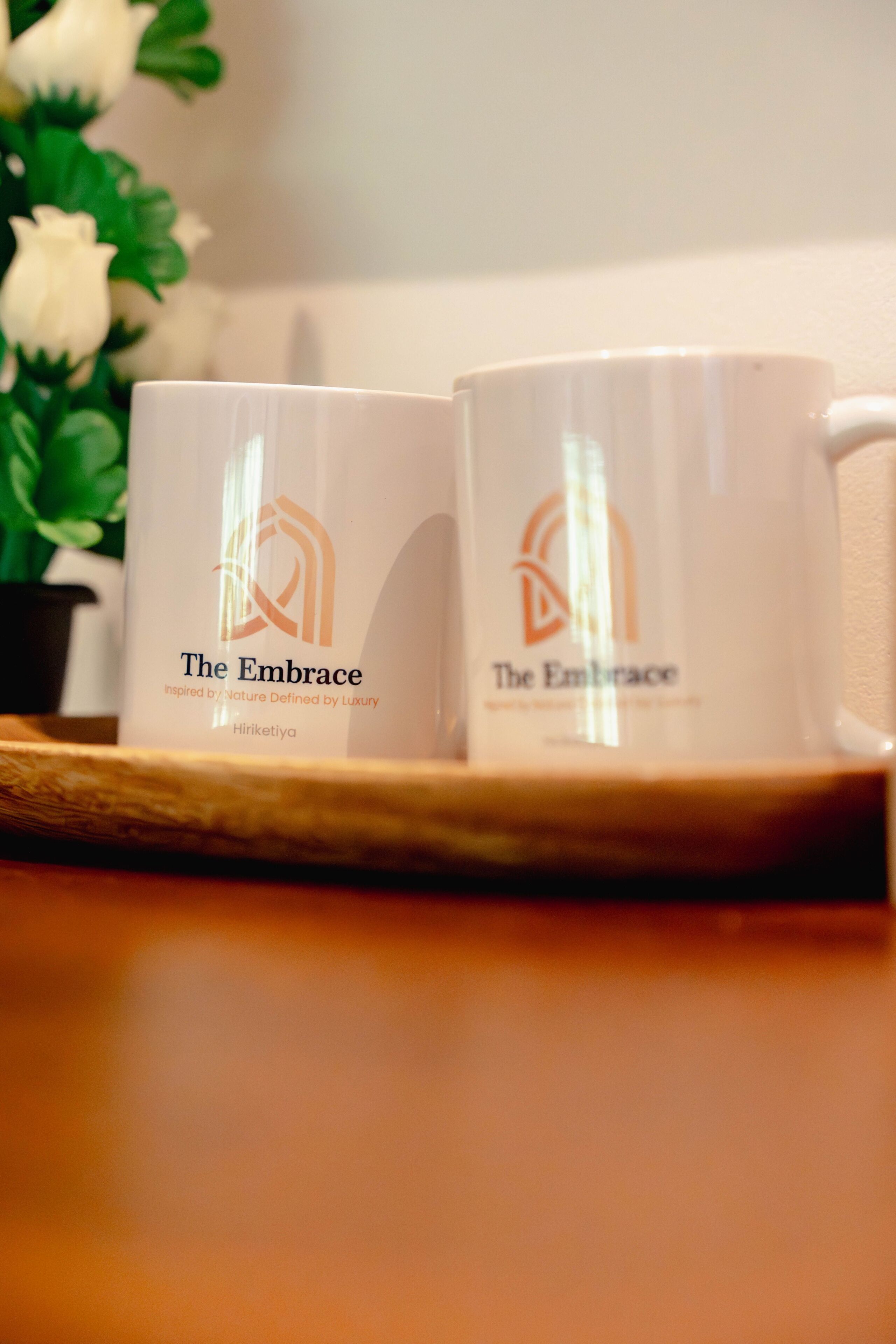 Twee witte mokken met het logo 'The Embrace' op een houten dienblad.