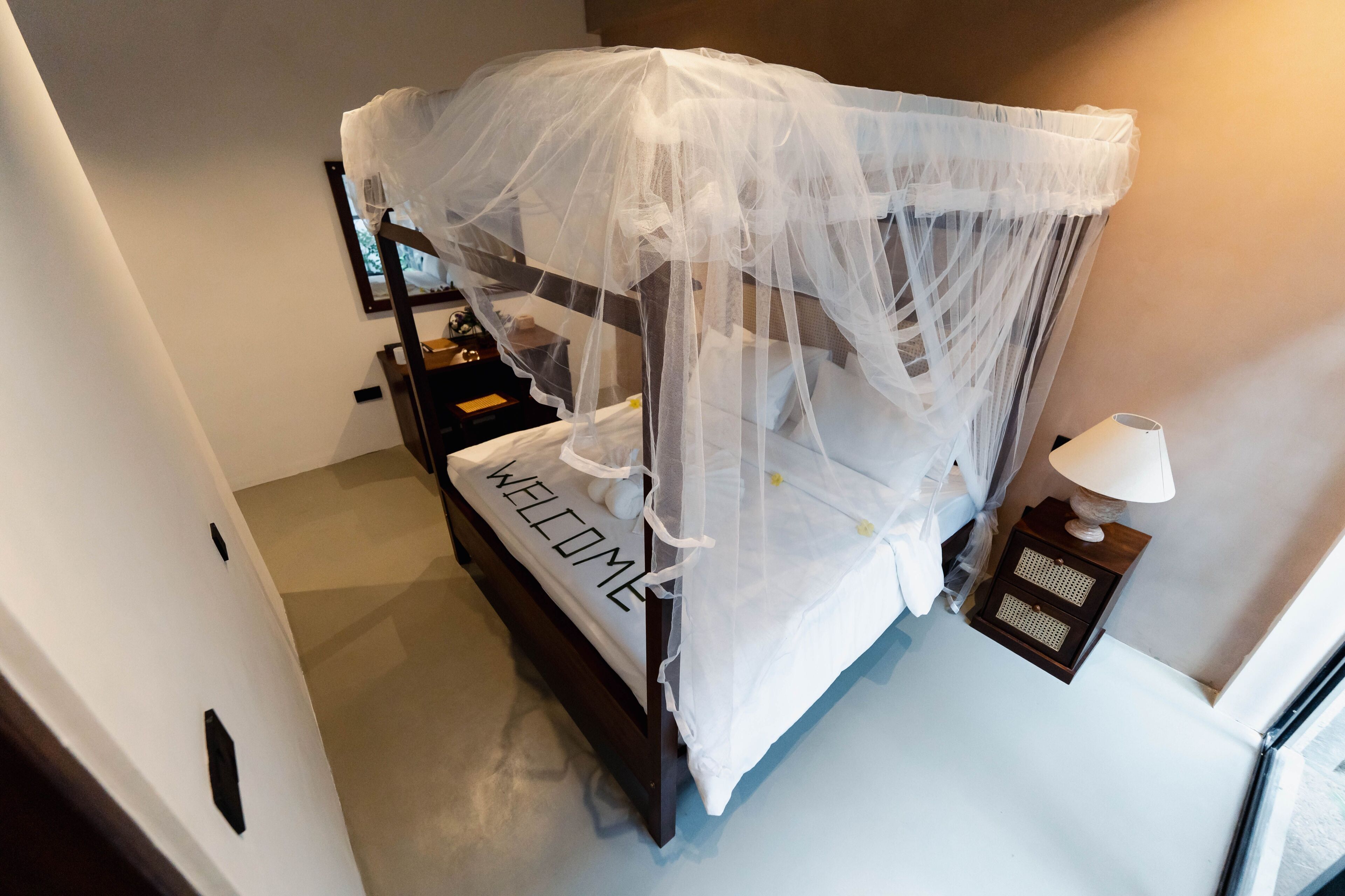 A bed with a canopy, a bedside table with a lamp, and a 'WELCOME' sign on the bed.